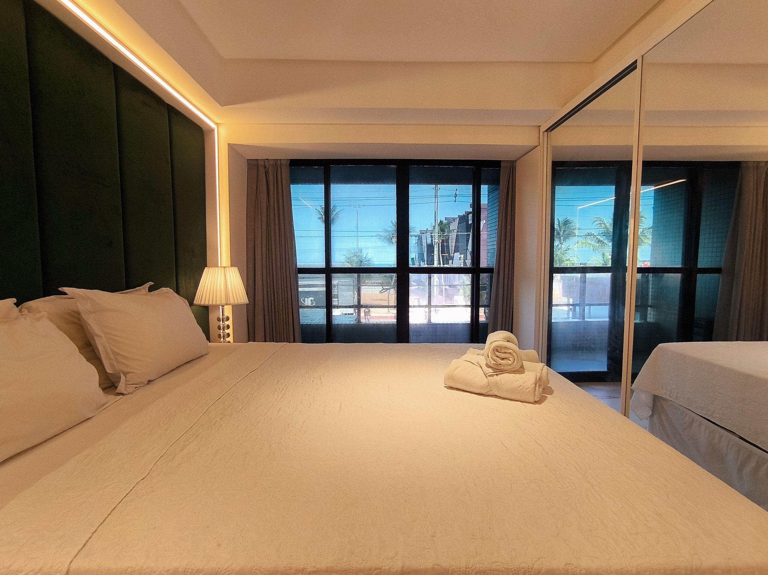 A hotel room with a large bed, a bedside table with a lamp, a window with a view of buildings, and a mirror on the wall.