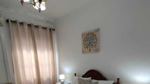 Ein ordentlich bezogenes Bett mit weißer Tagesdecke und zwei Kissen, ein Nachttisch mit Lampe, ein Sessel und ein Fenster mit Vorhängen.
