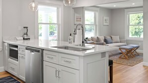 A modern kitchen with white cabinetry, a built-in dishwasher, and a large island with a sink. There are pendant lights above the island, and the kitchen opens to a living area with a sofa and a rug. - 2 Bed, 1.5 Bath Stunning Apartment, Central Andover Location (Andover)