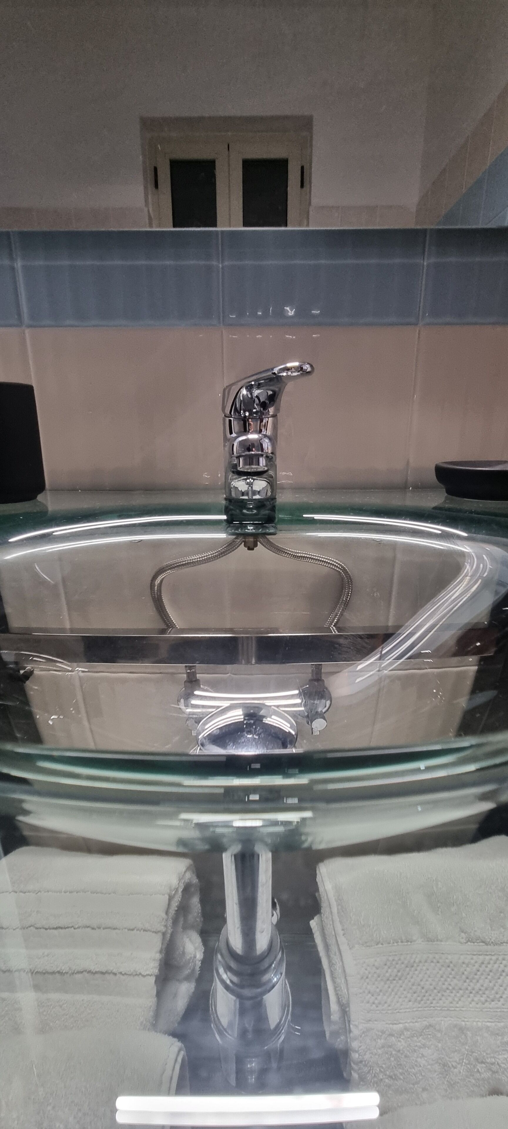 Ein durchsichtiges Waschbecken mit Glasabdeckung und chromfarbenem Wasserhahn, vor einer gefliesten Wand mit einem Fenster darüber.