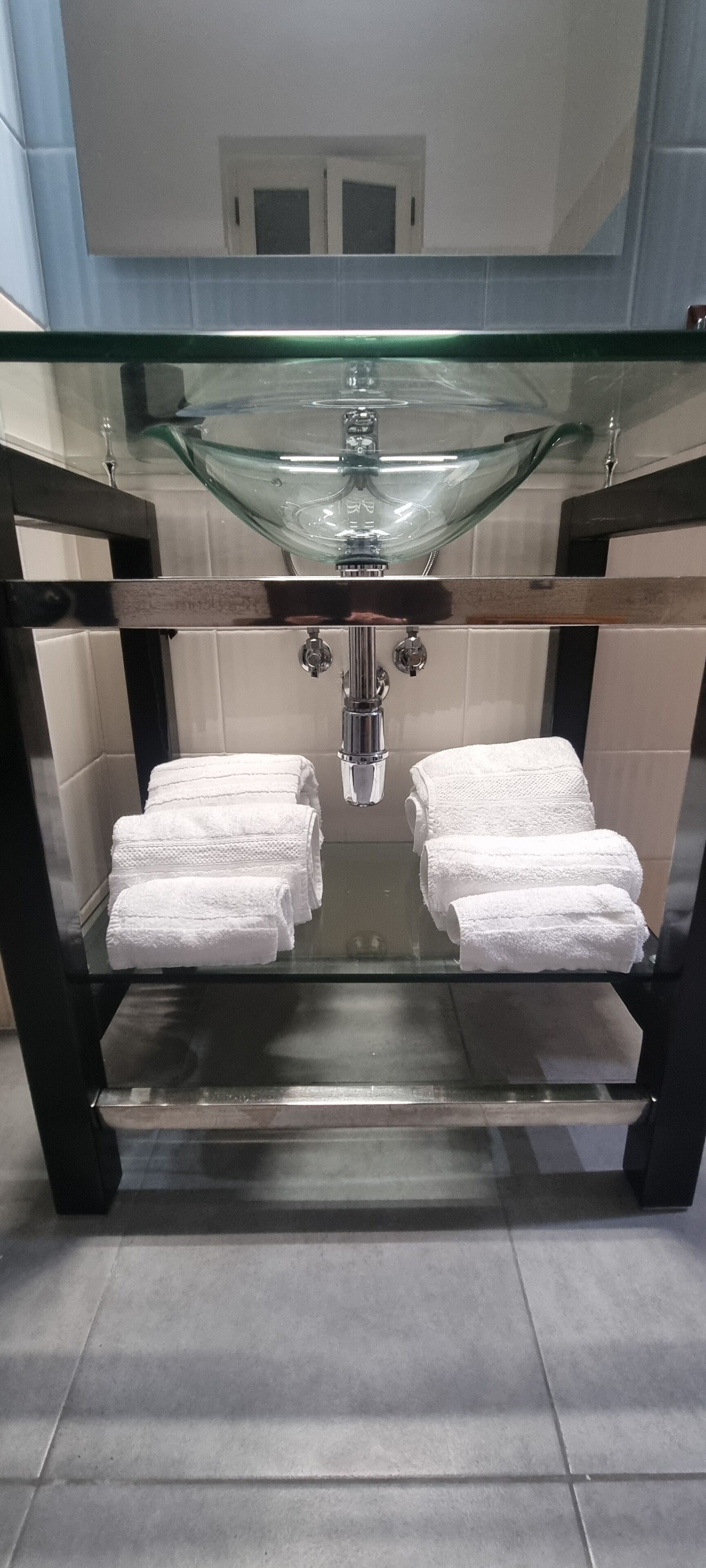 Un bagno con ripiano in vetro, asciugamani bianchi, lavandino in vetro e uno specchio.