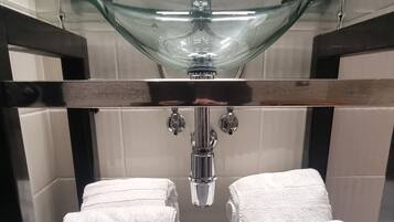 Un bagno con ripiano in vetro, asciugamani bianchi, lavandino in vetro e uno specchio.