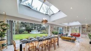 A spacious dining area with a long table set for a meal, surrounded by chairs, and a view of a garden and house through large windows. - Legoland Basecamp | Perfect for Families | Sleeps 15 (Windsor)
