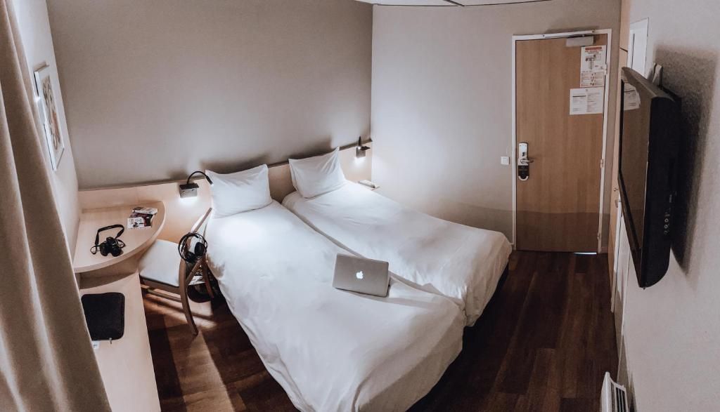 Une petite chambre d’hôtel avec un lit simple, une table de chevet avec une lampe, un ordinateur portable et un téléviseur fixé au mur.