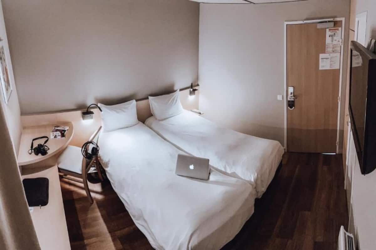 A small hotel room with a single bed, a bedside table with a lamp, a laptop, and a TV mounted on the wall.
