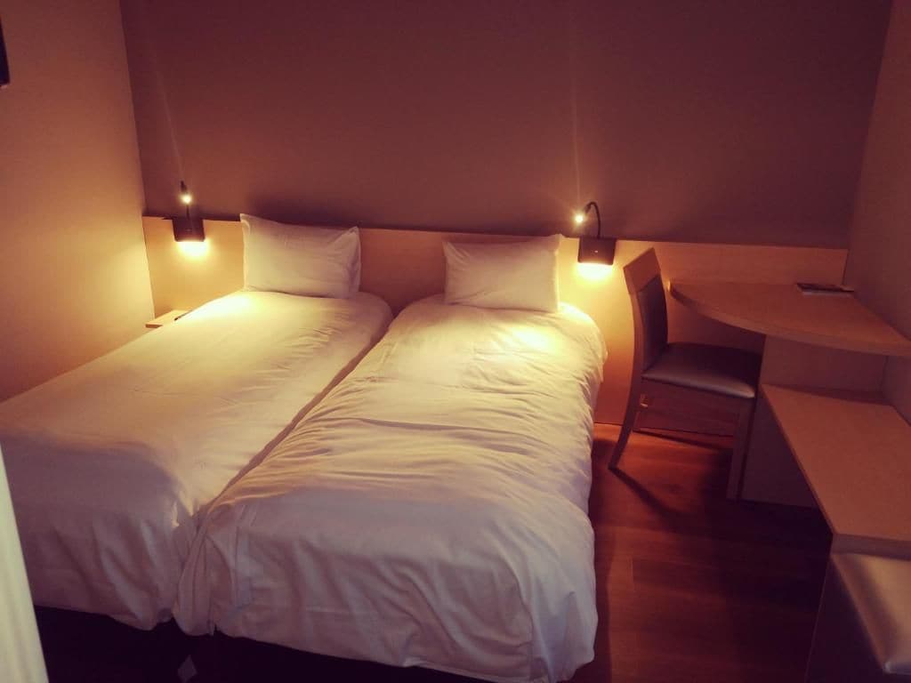 A double bed with white linens, two bedside tables with lamps, a small desk, and a chair.