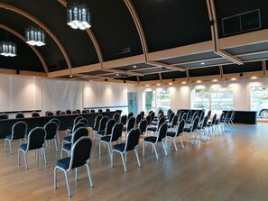 Une salle de conférence avec des rangées de chaises, une scène et de grandes fenêtres.