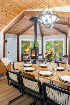 Open living and dining space with vaulted wood ceiling, fireplace & tree views.