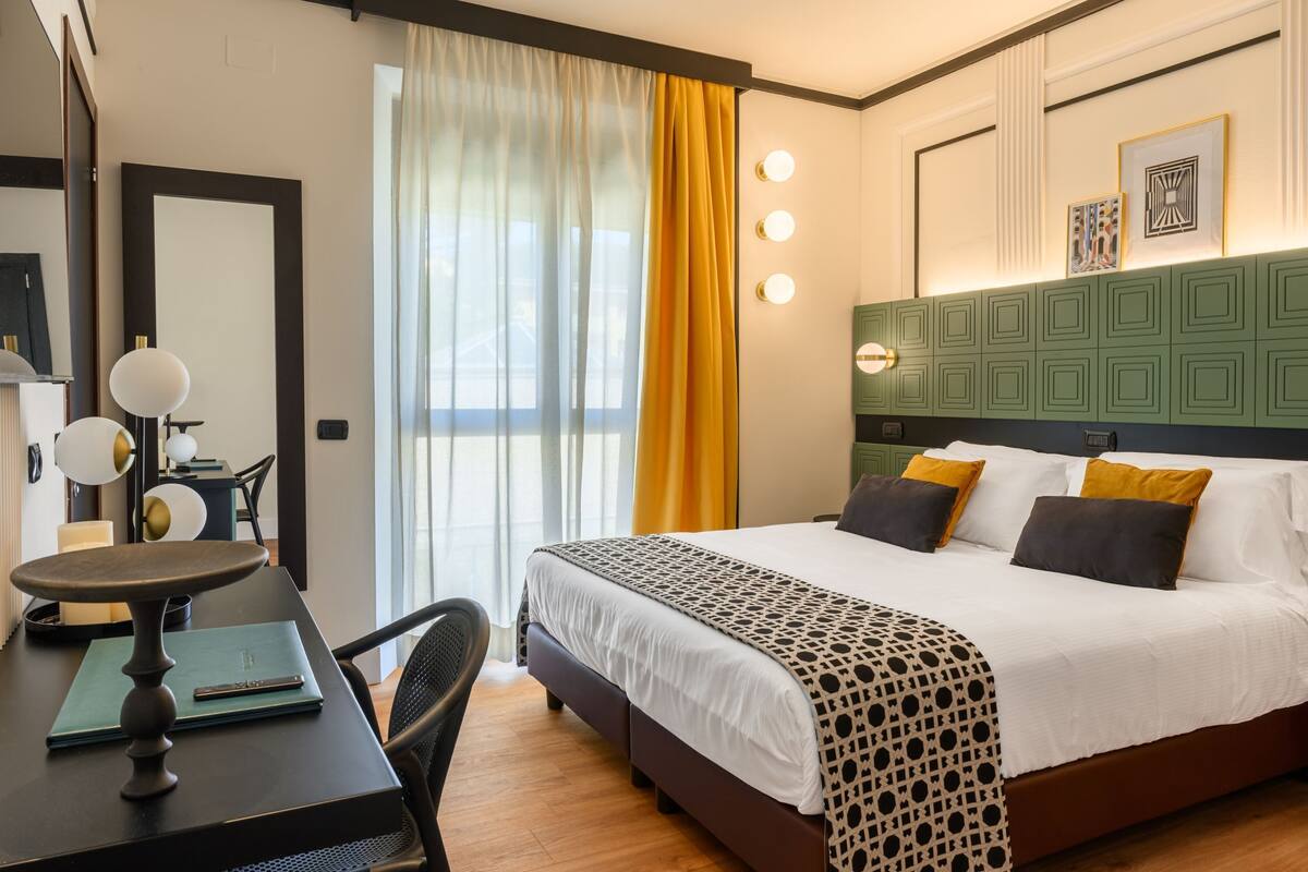Une chambre d’hôtel avec un lit, un bureau avec une lampe, une chaise et une fenêtre avec des rideaux.
