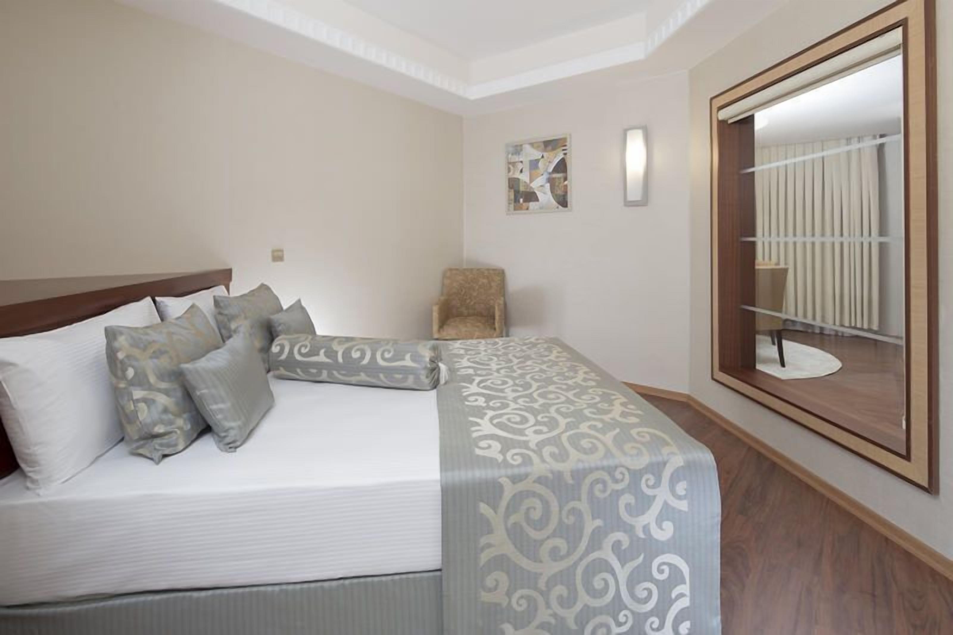 A neatly made bed with a patterned bedspread, a wooden headboard, and several pillows. There is a chair against the wall and a large mirror reflecting a room with a radiator.