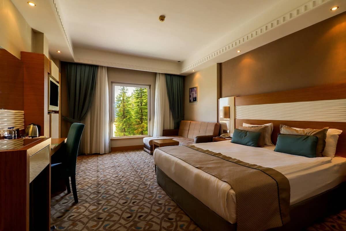 Ein Hotelzimmer mit einem großen Bett, einem Schreibtisch und einem Stuhl. Ein Fenster mit Vorh ängen bietet Blick auf Bäume.