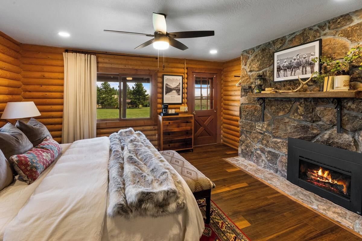 A log cabin room with a bed, a fireplace, a desk, and a view of the outdoors.