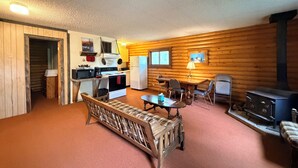 A cozy cabin interior with a wooden dining table, a sofa, a kitchen area with appliances, and a fireplace.