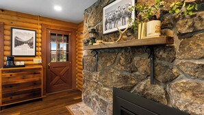 A log cabin interior with a stone fireplace, wooden shelves with books and a vase, and a door with a glass panel.