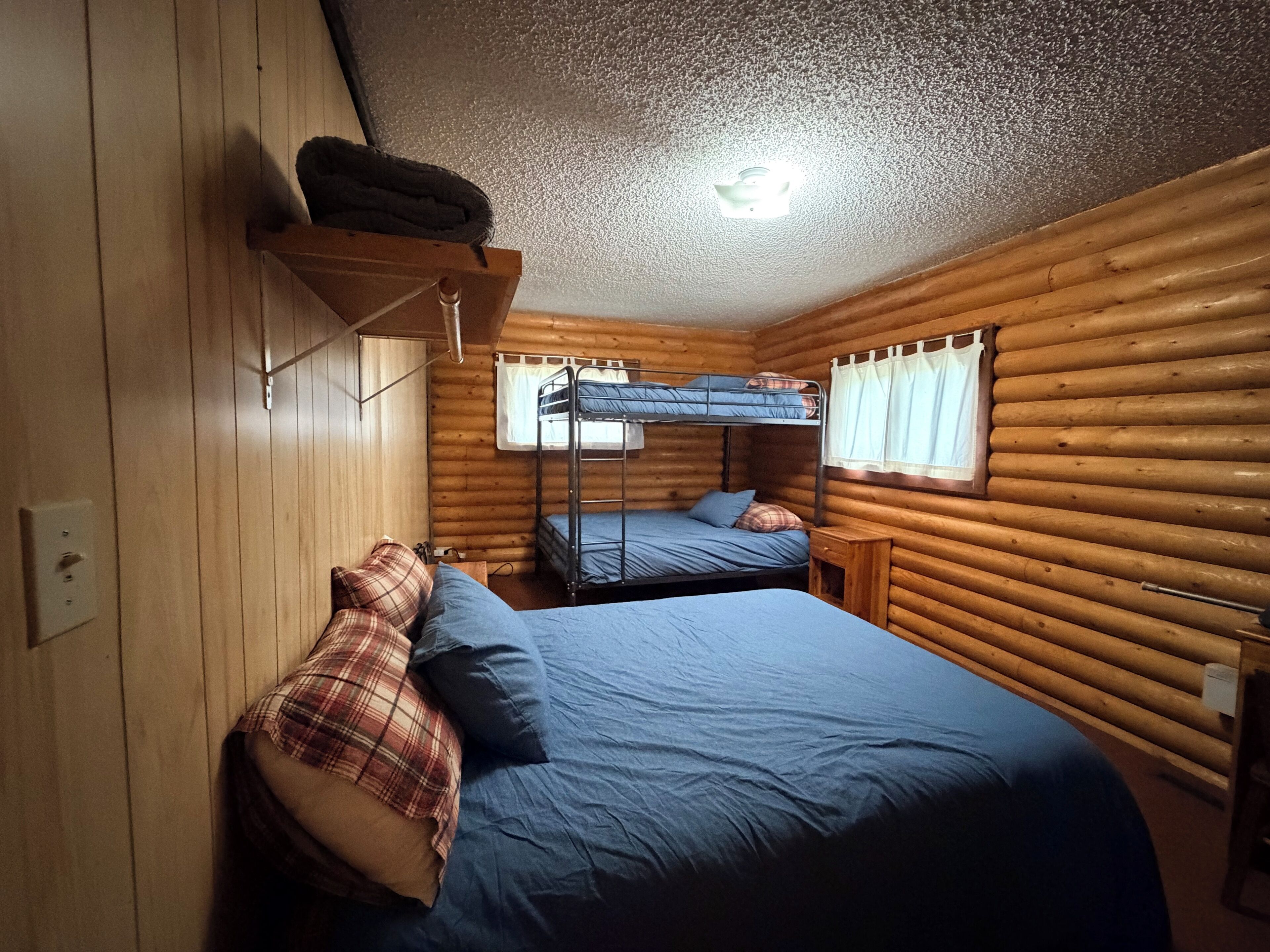 A cabin with a bunk bed, a single bed, wooden walls, a ceiling light, and a window with curtains.