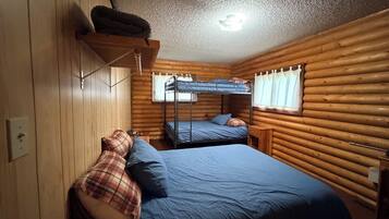 A cabin with a bunk bed, a single bed, wooden walls, a ceiling light, and a window with curtains.