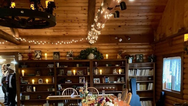 A cozy wooden cabin interior with a dining table set for six, surrounded by bookshelves filled with books and framed photos.