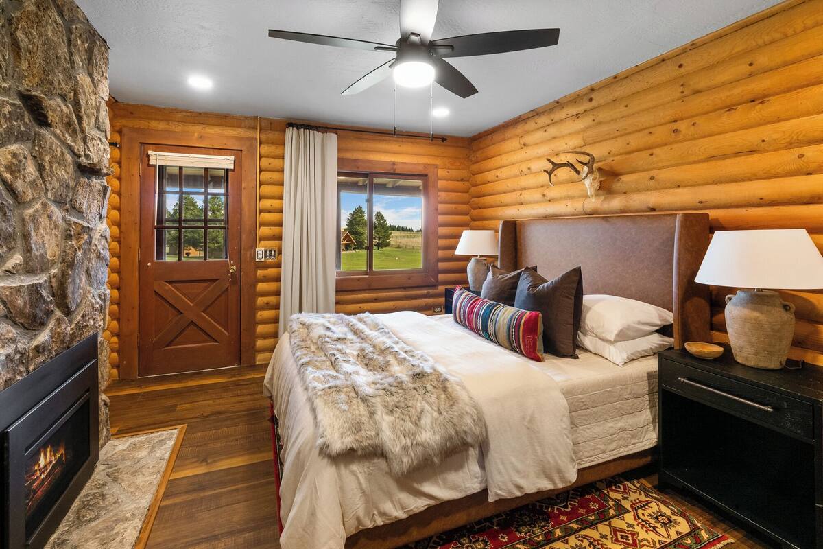 A log cabin bedroom with a stone wall, a fireplace, a bed with a fur throw, and a view of a green landscape.