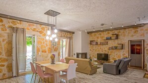 A spacious living area with a wooden dining table, pink chairs, a beige sofa, and a gray recliner. There is a fireplace and a bookshelf against the stone wall. - Villa Yiarios in Corfu (Corfu)