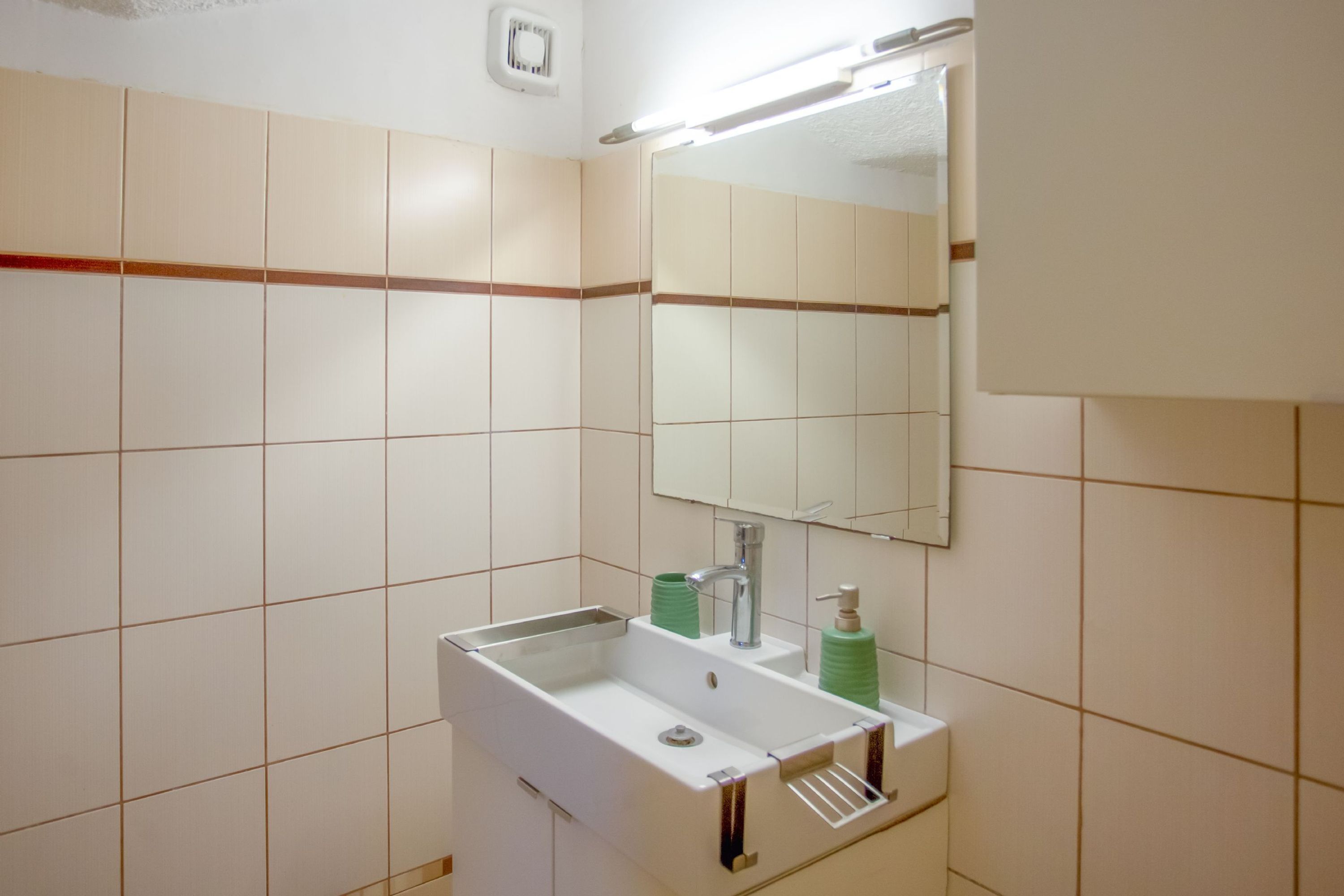 Un baño con un lavamanos blanco, dispensador de jabón verde y un espejo encima.