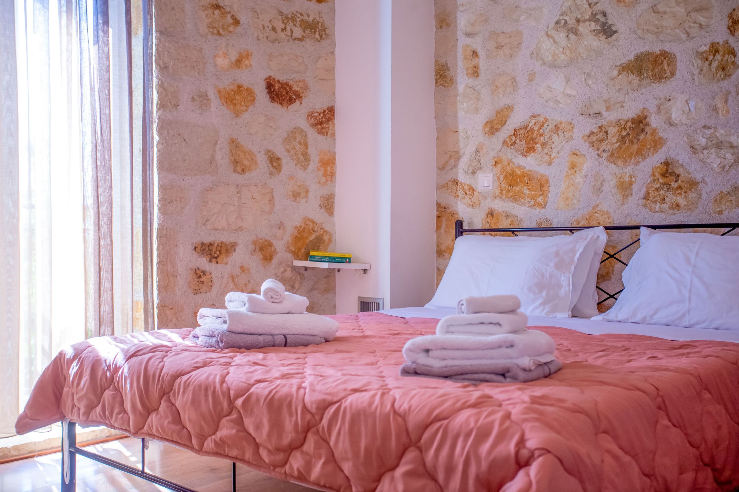 A hotel room with a stone wall, a bed with pink bedding, and towels neatly stacked on the bed.