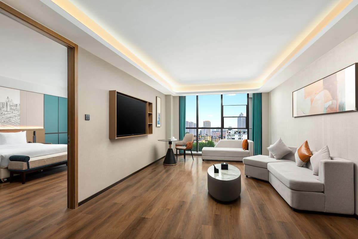A modern hotel room with a large bed, a sofa, a coffee table, and a city view through large windows.