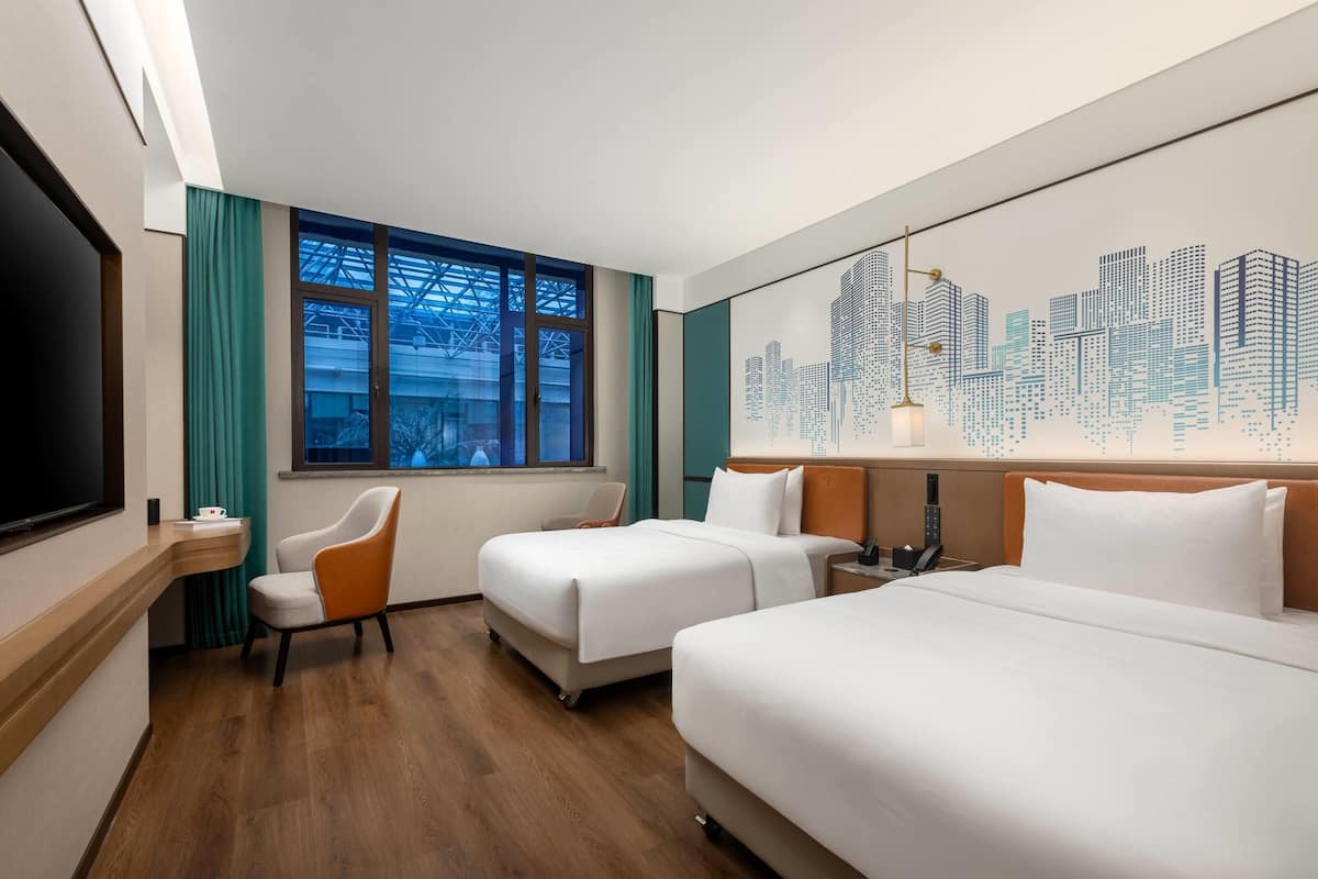 A hotel room with two beds, a desk, a chair, a cityscape mural, and a large window with curtains.
