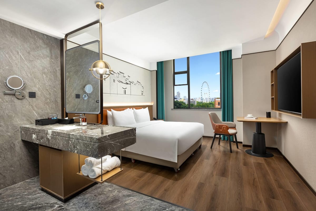 A hotel room with a bed, a vanity with a mirror, a desk, and a view of a cityscape.