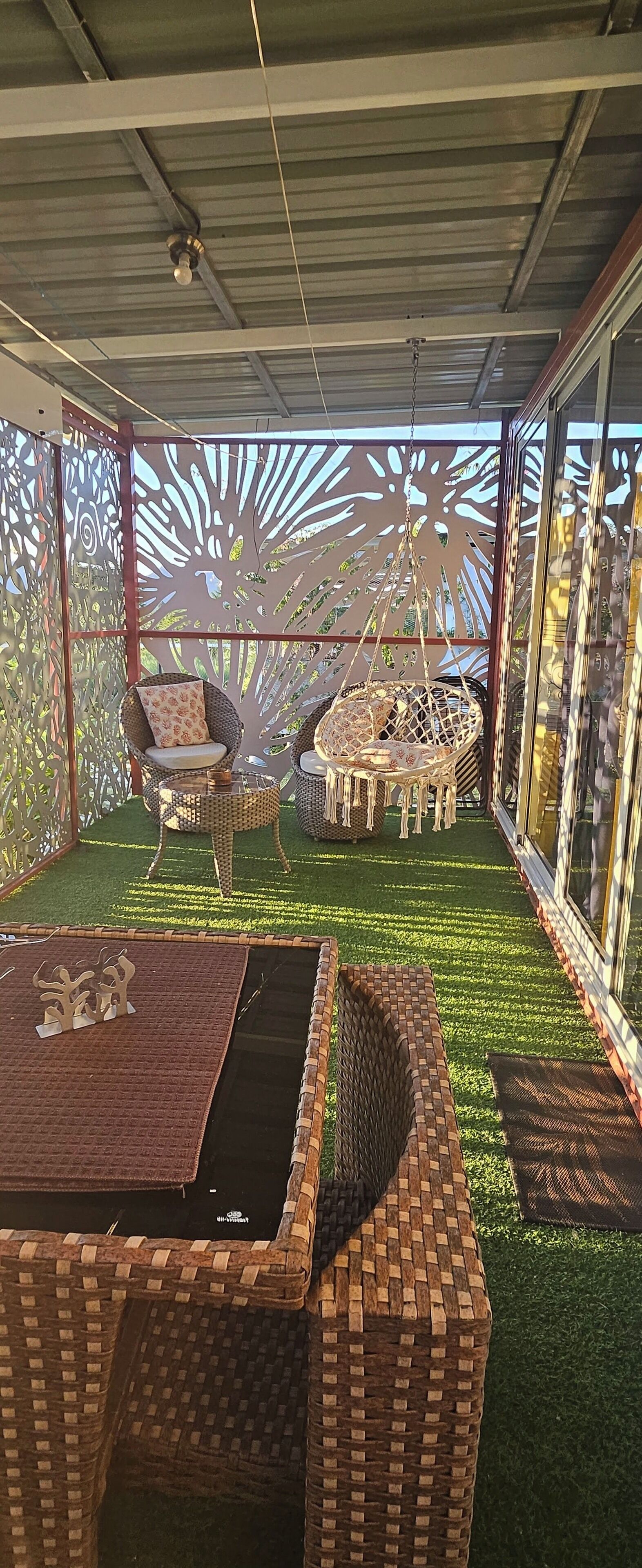 A covered outdoor seating area with wicker furniture, a hanging chair, and a decorative wall with cut-out patterns.