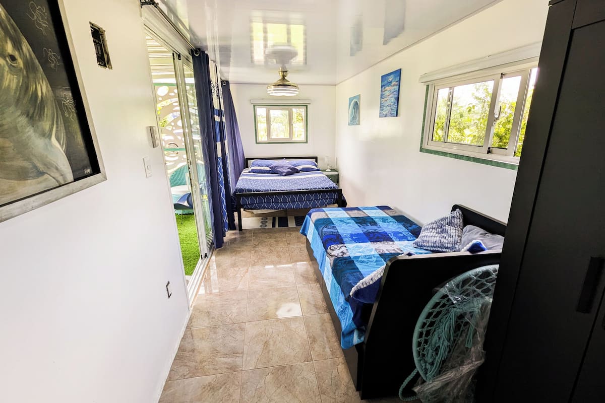 A narrow corridor leading to two bedrooms with blue bedding, a ceiling fan, and a window with a view of greenery.