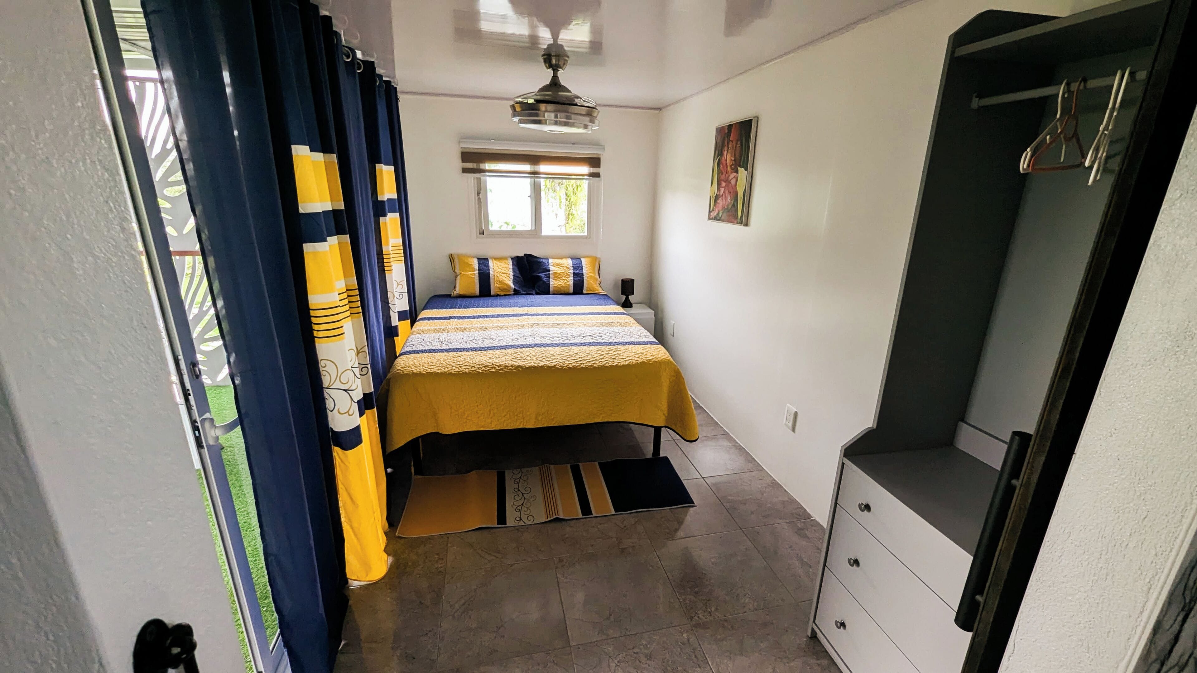 Une chambre à coucher avec un lit, des rideaux, un ventilateur de plafond, une fenêtre et une commode.