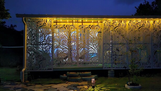 Un cancello metallico decorativo con motivi intricati, illuminato da luci a stringa, situato in un giardino con vialetto in pietra e piante in vaso.