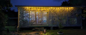 Una valla metálica decorativa con diseños intrincados, iluminada por luces colgantes, ubicada en un jardín con un camino de piedra y plantas en macetas.