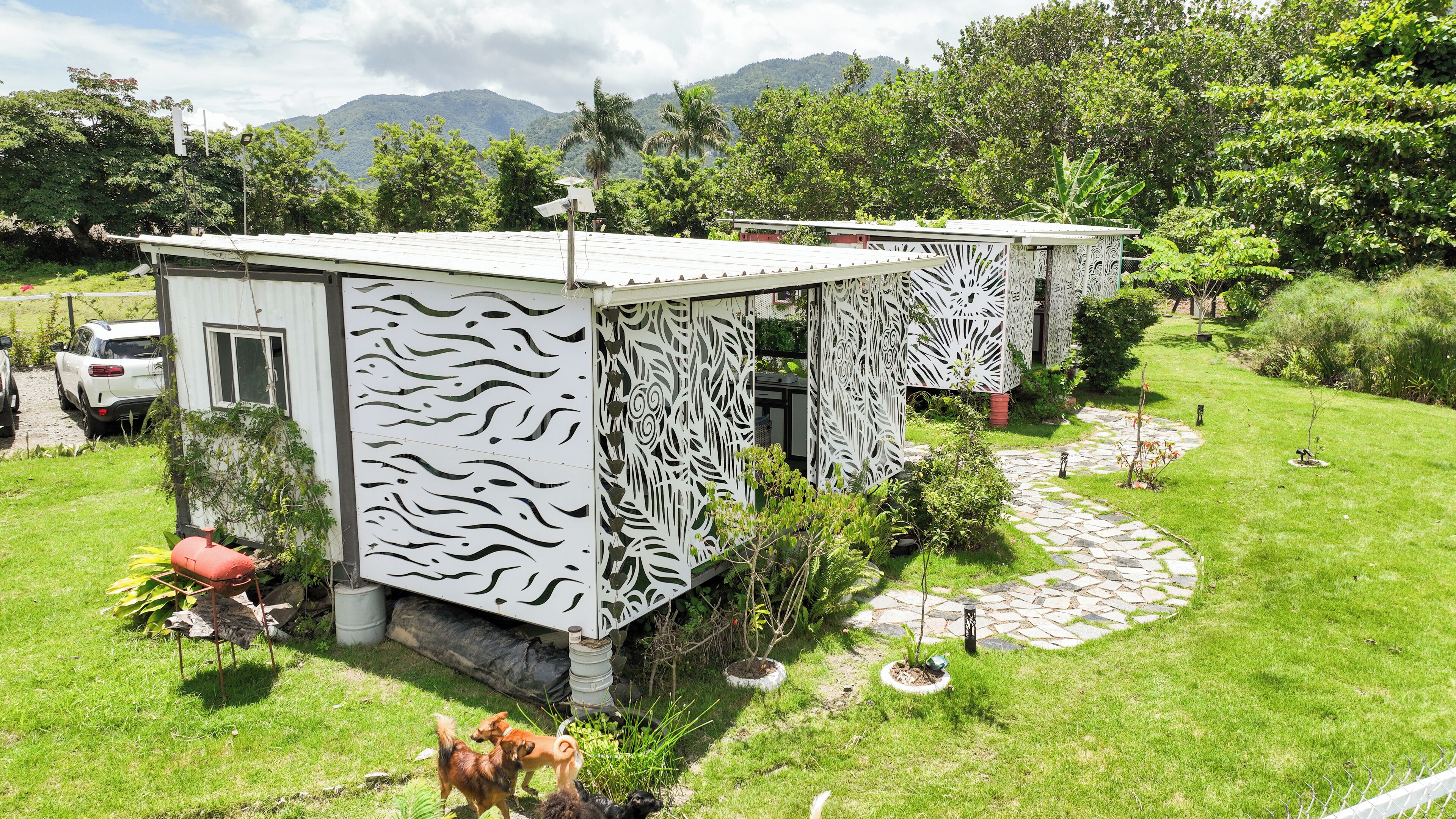 A small, modern house with a unique white facade and patterned design, surrounded by a well-maintained garden and a mountainous backdrop.