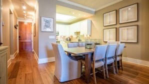 Un comedor con una mesa de madera y seis sillas, una cocina al fondo y cuadros en las paredes.