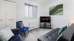 Ein modernes Wohnzimmer mit einer Couch, einem blauen Sessel, einem Fernseher, der 'NETFLIX' zeigt, und einer Heizung.