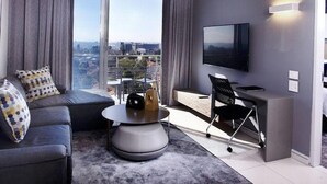 Ein modernes Wohnzimmer mit einer Couch, einem Couchtisch und einem Schreibtisch mit Stuhl. Ein Flachbildfernseher ist an der Wand befestigt, und ein großes Fenster bietet Blick auf die Stadt.