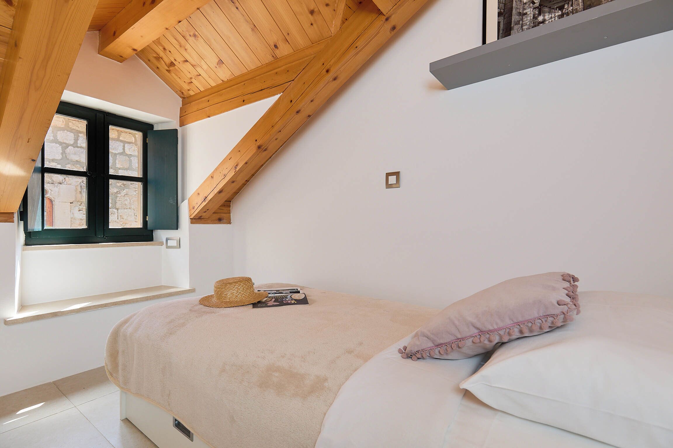 Een slaapkamer met een schuin dak, een bed met een beige deken, een strohoed en een tijdschrift op de vensterbank.