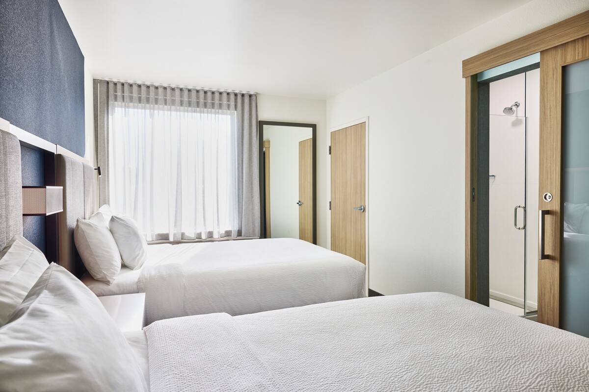A hotel room with two beds, a large window with curtains, a wooden wardrobe, and a shower area.