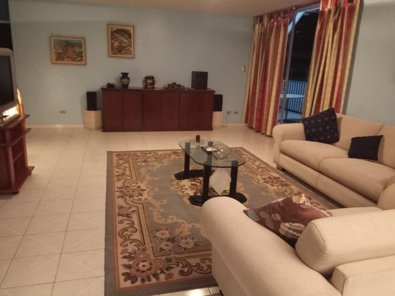 Ein Wohnzimmer mit einer Couch, einem Glastisch, einem gemusterten Teppich, einer Holzkommode und einem Fernseher.
