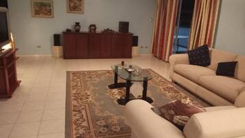 Ein Wohnzimmer mit einer Couch, einem Glastisch, einem gemusterten Teppich, einer Holzkommode und einem Fernseher.
