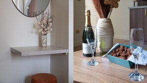 A room with a round mirror, a vase with dried flowers, a bottle of champagne, and a box of chocolates.