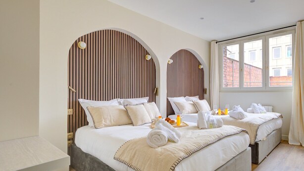A hotel room with two beds, a wooden slat headboard, a large window with curtains, and a breakfast tray on the beds.