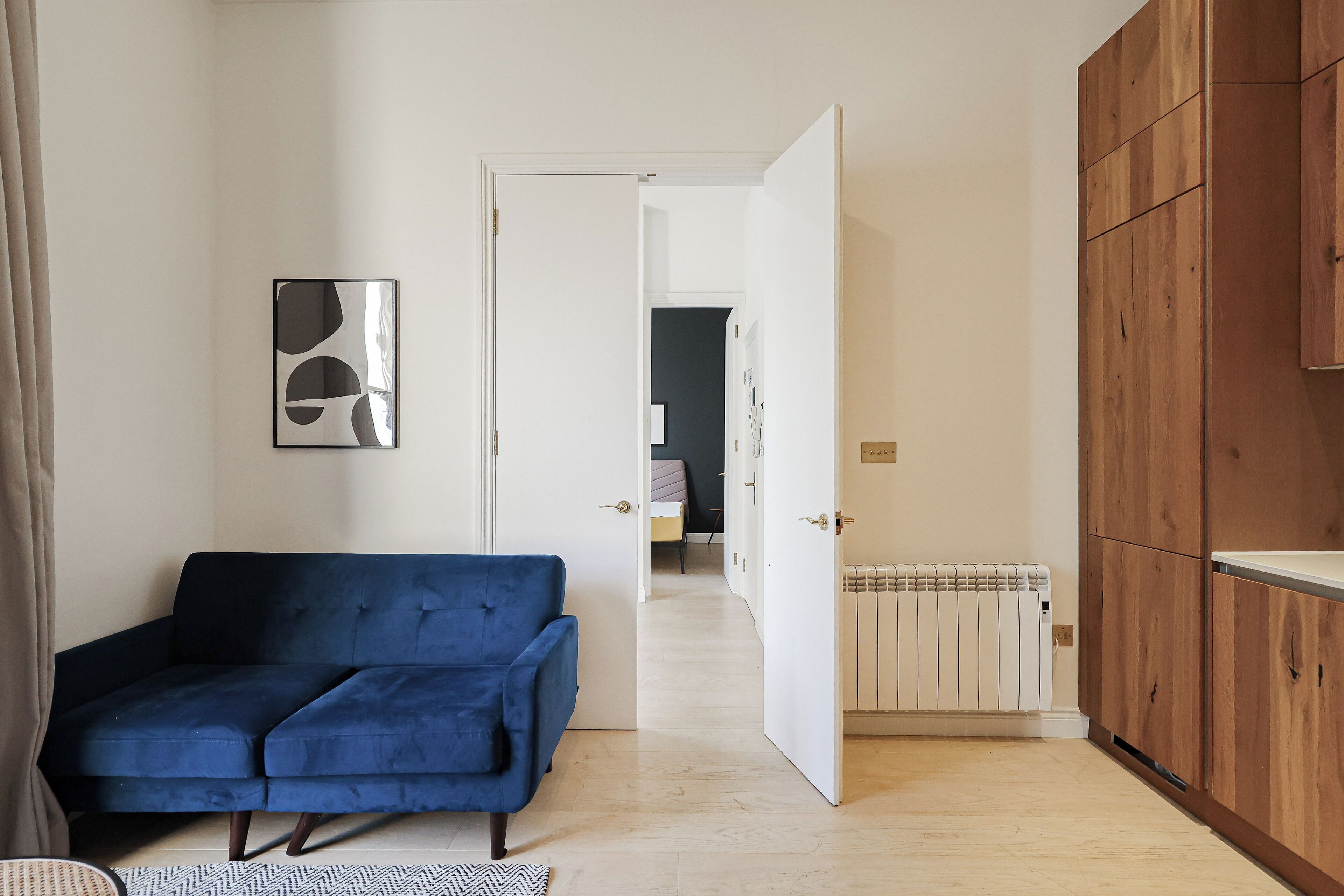 Una habitación con un sofá azul, un radiador, armarios de madera y una puerta que da a otra habitación.
