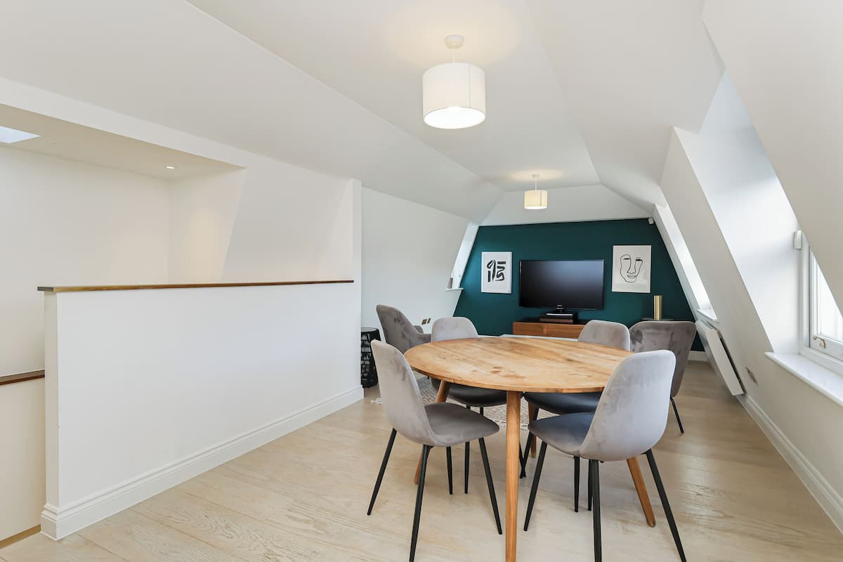 Un espace repas moderne avec une table en bois et des chaises à revêtement gris, un téléviseur à écran plat fixé au mur et un escalier menant à un niveau supérieur.