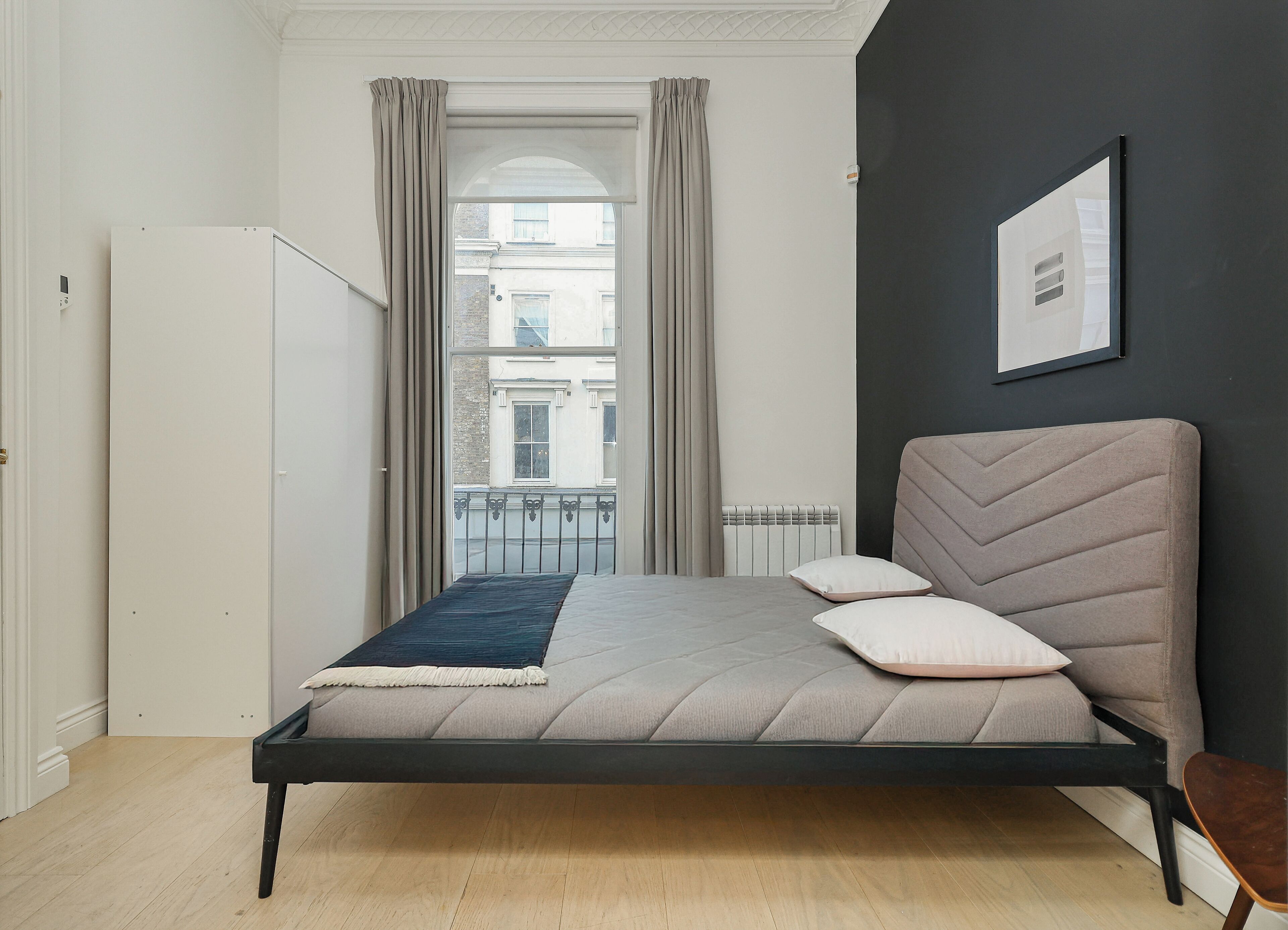 A bedroom with a bed, a radiator, a window with curtains, and a view of buildings outside.