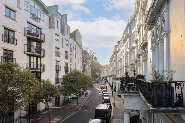 A street view of a residential area with multi-story buildings, parked cars, and trees lining the street. - Kensington Gardens Apartments (London)
