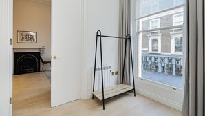 Een kamer met een houten vloer, een zwart metalen schap, een witte deur, een open haard en uitzicht op een gebouw door een raam.