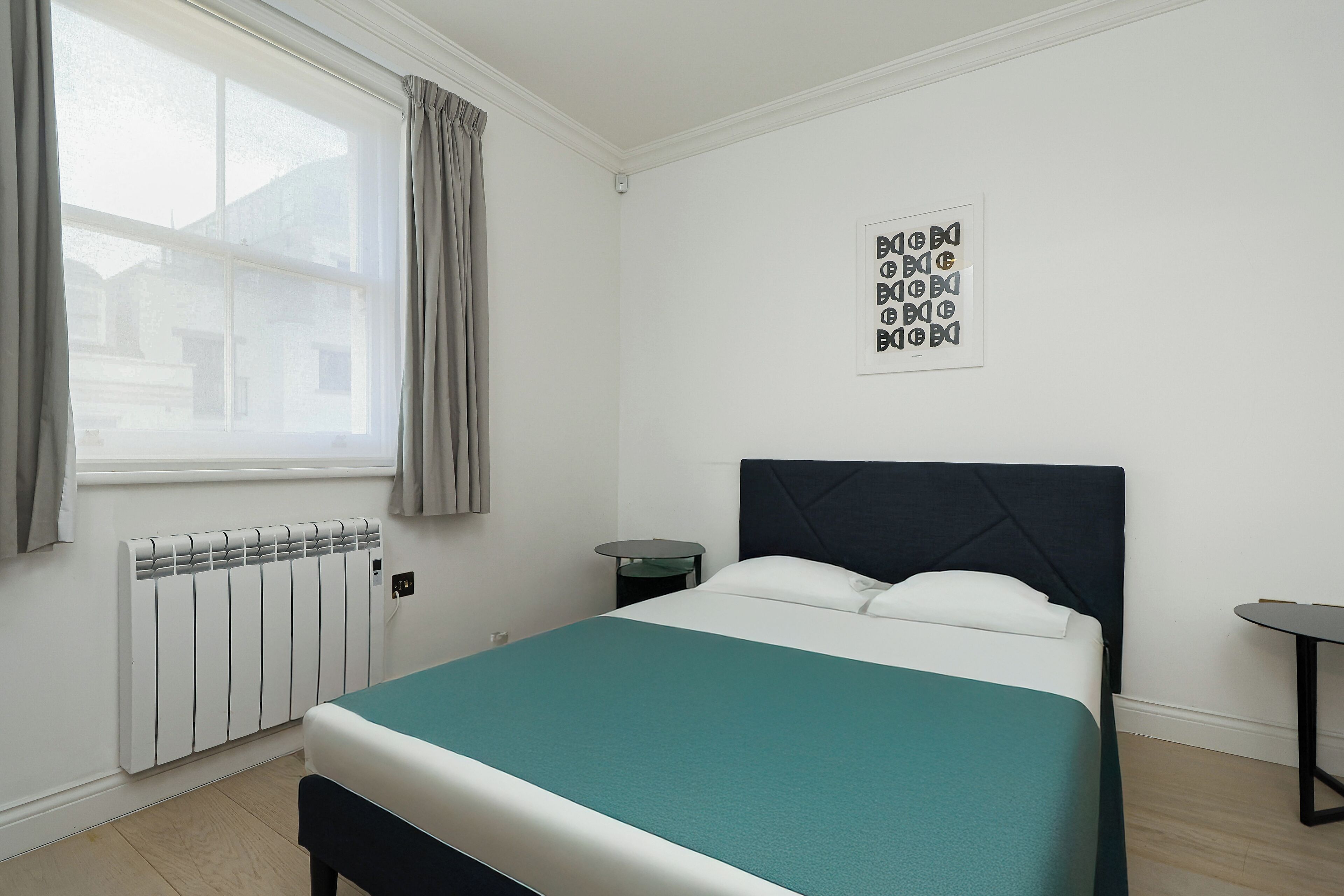 Ein Schlafzimmer mit Bett, einem Fenster mit Vorhängen, einem Heizkörper, einem Nachttisch und einem Wandbild.