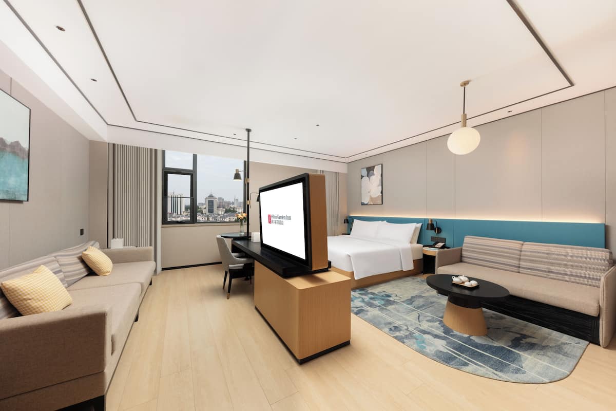 A modern hotel room with a large bed, a sofa, a desk with a flat-screen TV, and a city view through the window.